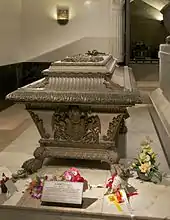 Photo d'un sarcophage clair avec ornements en bronze posé sur un dallage de marbre blanc.