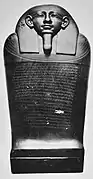 Black and white image of a dark stone coffin with a human face, the coffin stands upright facing the viewer.