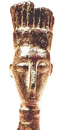 Statuette dorée sur fond blanc d'un visage en lame de couteau avec une coiffe haute.