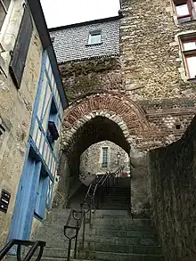 Vue d'une porte dans une muraille, avec des bâtiments situés à gauche et à droite, dont l'arc est constitué de briques et traversée par un escalier avec une forte pente
