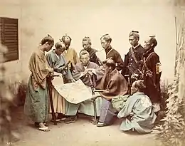 Photo colorisée de dix hommes en tenues de samouraï, trois assis au premier plan, sept debout au second, sur fond sépia clair.