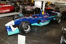Photographie d'une monoplace de Formule 1 bleu foncé et vert clair, vue de trios-quarts, dans un musée.