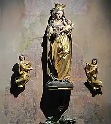 photographie en couleur montrant une statue dorée d’une femme tenant un enfant, avec de chaque côté des anges dorés, le tout devant un mur peint d’une fresque rouge avec des personnages en prière