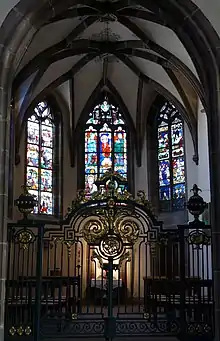 photographie en couleur montrant une petite chapelle voûtée, avec à l’avant plan une grande grille en fer forgé avec des dorures et à l’arrière-plan trois baies dotées de vitraux colorés