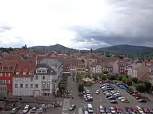 Vue plongeante d'une ville avec une grande place servant de parking, un manège jaune, une église. En arrière plan, des basses montagnes.