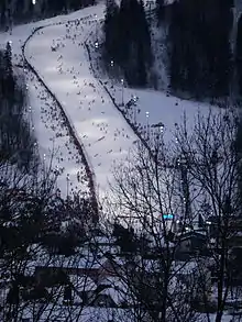 Vue de haut et lointaine d'une piste de skis avec des skieurs, un village apparaissant au premier plan.