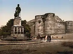 Photo d'une place pavée avec une statue en son centre, le château d'Angers en arrière-plan; deux petits attelages contournent la statue.