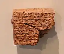 Tablette carrée en matériau rouge, cassée dans un angle, et inscrite de signes cunéiformes.