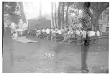 Photo en noir et blanc d'écoliers assis sur des bancs en plein air regardant un grand tableau.