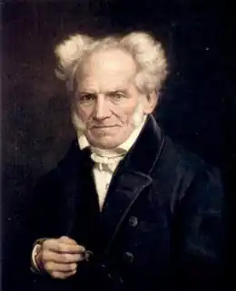 Portrait d'un homme portant un manteau noir, regardant droit devant lui, sans sourire, tenant sa main droite devant lui. Il est chauve, et les cheveux blancs sur les côtés de sa tête sont orientés vers l'extérieur.