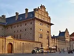 L'imposante façade à pignons et sgraffites d'un palais, de couleur grise mais éclairée par le soleil couchant, avec un train touristique en stationnement.