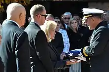 Une femme, au milieu, reçoit un drapeau américain des mains d'un officier en uniforme. Deux hommes à l'avant-plan, foule à l'arrière plan.