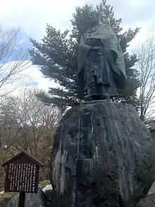 Photo couleur montrant, sur fond de ciel bleu nuageux, une statue en pied d'un homme en tenue de moine sur un rocher gravé d'une inscription.