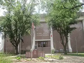 Vue d'un bâtiment à la façade en marbre marron. Devant sont présents deux arbres, un monument et un mat avec le drapeau américain.