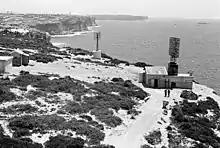 Photographie noir et blanc de deux antennes (une sur un bâtiment) avec des falaises maritimes à l'arrière plan.