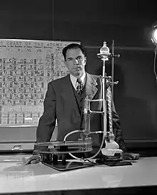 Photographie en noir et blanc de Glenn Theodore Seaborg de face derrière un montage de chimie avec un tableau périodique en arrière-plan.