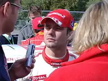 Portrait de Sébastien Loeb avec une casquette répondant à des journalistes tendant un dictaphone.