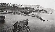 Photographie prise depuis un navire montrant une forteresse en ruine surplombant une pente escarpée menant à une étroite plage.
