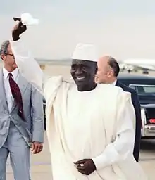 photo en buste d'un homme vêtu d'un boubou et d'une toque de couleur blanche, saluant de la main, sur un tarmac d'aéroport
