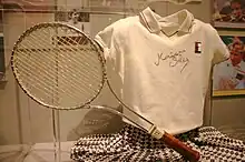 Photo des tenue et raquette de Seles à son retour en 1995. Polo blanc dédicacé, jupe à carreaux bleu marine et blanc.