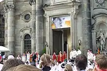 Photo de la façade d'un cathédrale avec plusieurs prélats, la foule et une grande photo d'un homme jeune