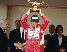Photo d'Ayrton Senna de face, plan américain, brandissant le trophée de la victoire. En arrière-plan, les officiels dont le Prince Rainier de Monaco.
