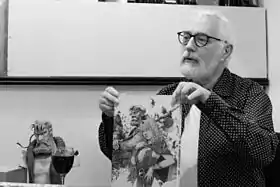 Serge Le Tendre présentant la couverture du tome 1 de l'intégrale de la série, représentant les personnages de Bragon et Pélisse. Un buste en résine de Bragon se trouve à ses côtés.