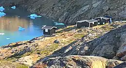 Photographie en couleurs de bâtiments installés sur les rives d'un fjord, des roches aux formes arrondies visibles au premier plan.