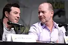 Photo de deux hommes assis derrière des tables et devant des microphones.