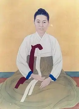 Peinture d'une femme, assise en tailleur de face. Une large robe commence au-dessus du ventre et couvre intégralement le bas du corps.