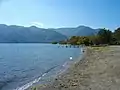 Photo couleur montrant les eaux bleues d'un lac, à gauche, une étendue de sable qui se termine, au second plan, par un bateau devant une forêt, à droite. En arrière-plan : un massif montagneux recouvert de forêts vertes sur fond de ciel bleu.