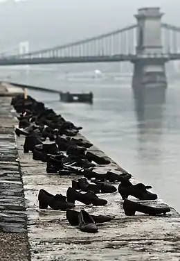 Photographie d'un quai au bord d'une rivière, sur lequel sont présentes plusieurs chaussures en métal au bord du quai ; au fond, un pont.