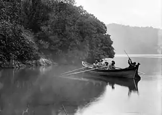 Photographie ancienne en noir et blanc d'une barque quittant un rivage dont les arbres surplombent directement la mer.