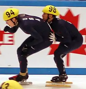 Un patineur américain en combinaison sombre en pousse un autre de derrière dans le cadre d'un passage de relais en patinant.