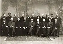 Photo en noir et blanc de 20 hommes posant en habit de cérémonie dans une salle richement décorée.