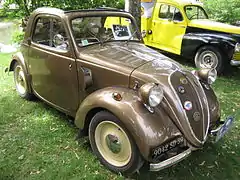 Photo d'une voiture de type coach, de couleur beige et de petite taille, stationnée dans l'herbe à côté d'un autre véhicule.