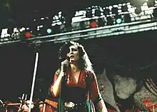 Une femme au teint blanchâtre et aux yeux très maquillés de noir, avec d'épais cheveux noirs ébouriffés, chante sur scène en tenant un micro.