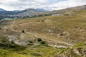 Alpage parsemené de trous et de bosses laissant deviner des ruines et, en arrière-plan, une station de ski.