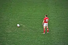 Photo d'une homme debout, de dos, immobile, s'apprêtant à frapper un ballon de rugby place quelques mètres devant lui.