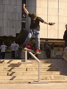 Dans une rue, un skater effectue un slide, il descend un petit escalier en équilibre sur la rambarde centrale. Il maintient l'équilibre avec ses bras, son skateboard est directement posé et glisse sur la rambarde.
