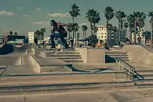 Photo d'un skater en train d'effectuer une figure de skate à Venice Beach. Il est pris en l'air durant un saut à 1 à 2 mètres de hauteur et son skate effectue des rotations en dessous de lui. Le décor est principalement composé d'un multitude d'escaliers en béton de quelques marches. En arrière-plan assez loin, il y a des skaters et beaucoup plus loin, on aperçoit des cocotiers, la plages et des immeubles.