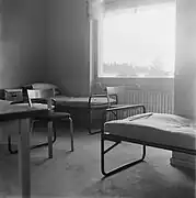 Photo en noir et blanc de l'intérieur d'une chambre. Elles ont des grandes fenêtres et un grand radiateur. Une table et une chaise sont visibles, ainsi que deux lits en métal sur lesquels sont posés des matelas fins.