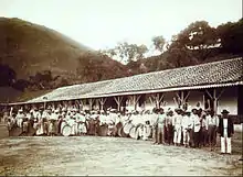 un important groupe d'hommes, de femmes et d'enfants tenant des instruments aratoires, debouts devant un long bâtiment bas avec des collines en arrière-plan