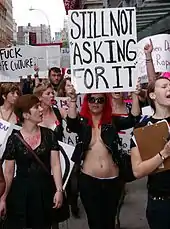 photographie couleur de plusieurs femmes manifestant. Une au centre porte la pancarte : Still not asking for it