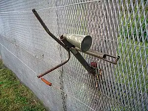 Engin de forme conique montée sur le côté d'une clôture métallique. Des fils de détente lui sont attachés et courent le long de la clôture.