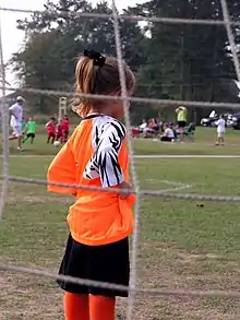 vue de derrière les filets d'une jeune gardienne de but de dos habillée en maillot orange et short noir.