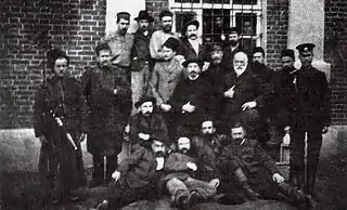 Photographie de 19 hommes, coiffés et moustachus, posant debout ou couchés devant un bâtiment avec une fenêtre grillée.
