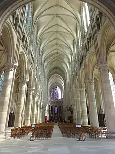 Une allée balisée de piliers, au plafond, presque au ciel, des croisées, au sol une multitude de chaises bien rangées, derrière les piliers des vitraux sans motifs.