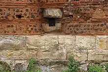 Vu d'un mur composé de plusieurs parties, d'un soubassement, d'une partie en briques et en petites pierres, avec au milieu un dispositif en pierre avec un trou.