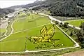 Photo couleur d'une vallée comprenant plusieurs rizières. Dans l'une d'elles, au premier plan, des plants de riz de différentes couleurs (vert, jaune et noir) dessinent un cheval ailé, au-dessus d'un message en coréen.